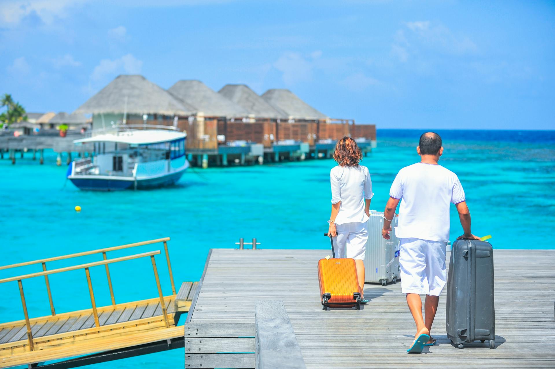 A couple holding briefcases walks on a wooden bridge by the sea during a beach vacation under a bright, sunny sky.