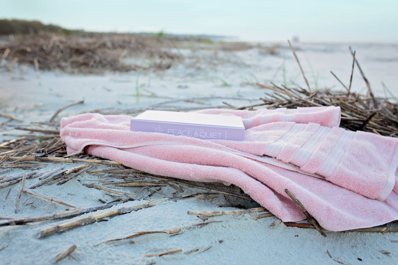 A pink beach towel spread on golden sand with a book placed on top, capturing the essence of beach essentials for relaxation.