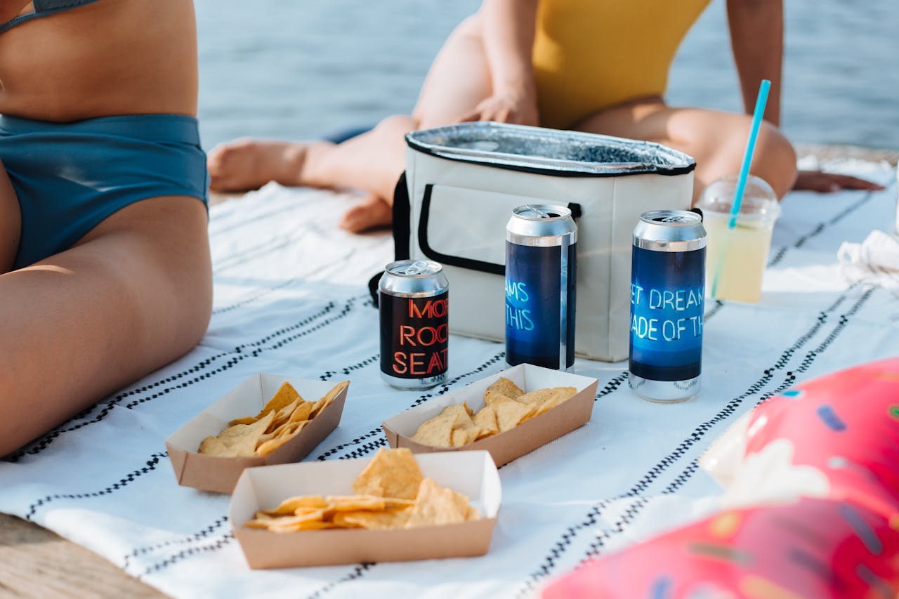 Cooler bag with cold drinks and chips on a cloth, with women enjoying their time at the beach, surrounded by beach essentials.