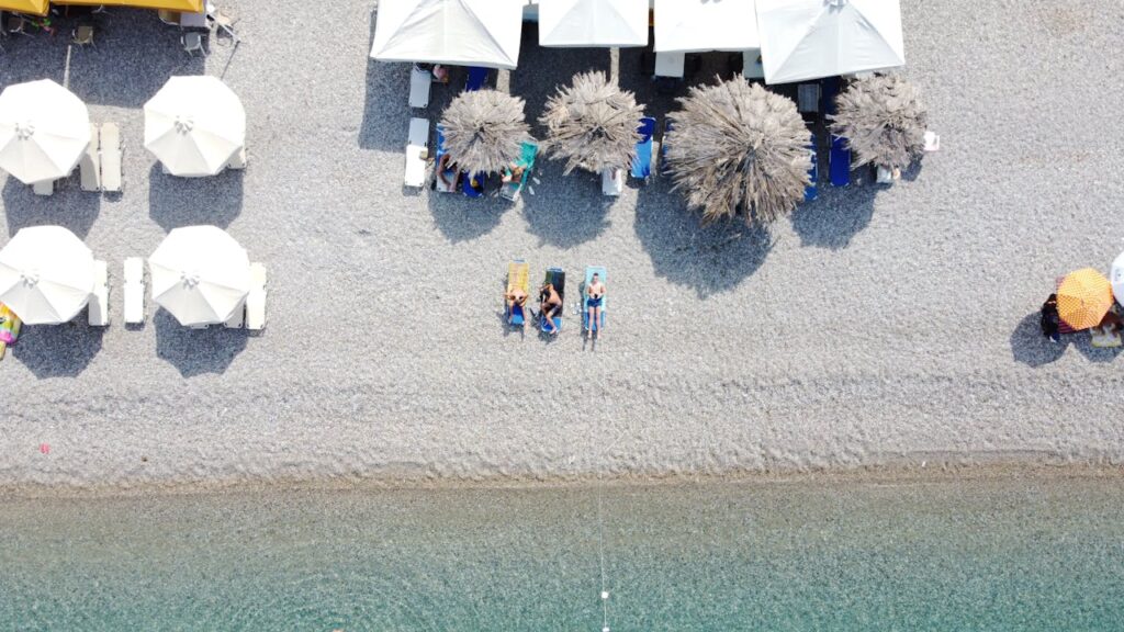 A stunning sky view of a beach with people sitting and enjoying the relaxing sea, surrounded by nearby umbrellas and shaded areas, featuring beach essentials.