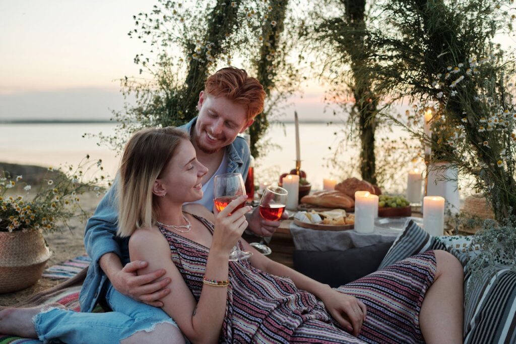 A couple enjoying drinks and pizza near a lake with a cozy setup, celebrating lovely day in a romantic evening setting.