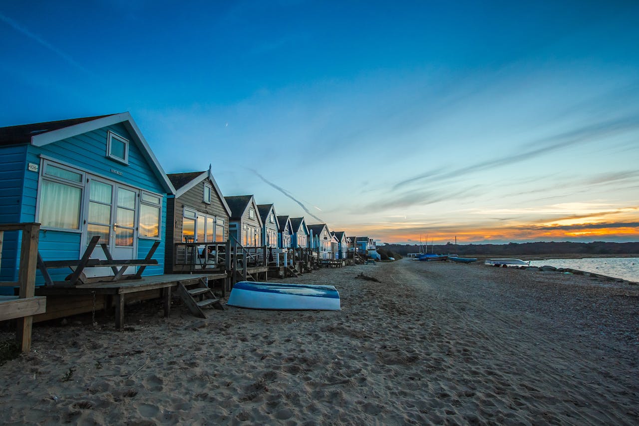 A row of charming wooden huts near the beach at sunset, showcasing a prime location—an important factor in how to become an Airbnb host. A boat rests nearby, adding to the scenic coastal vibe.